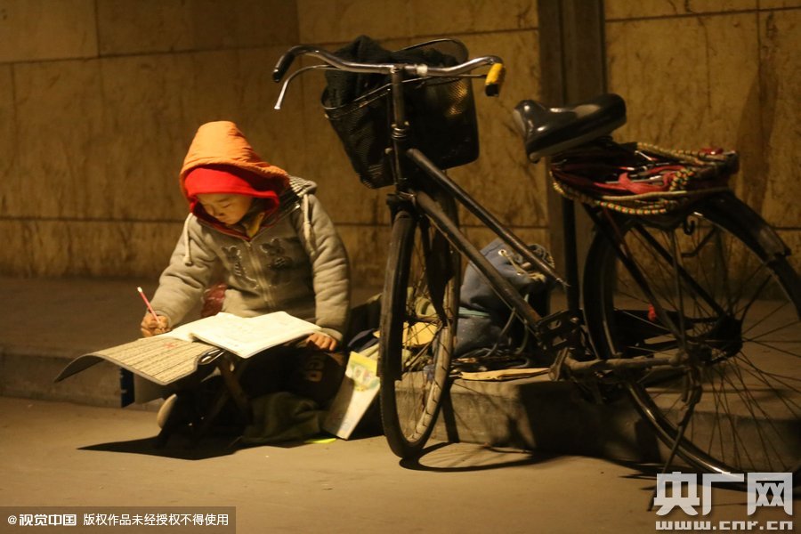 鄭州中原路京廣鐵路大橋下涵洞的馬路牙子邊，一位戴著紅色辣椒帽的小男孩趴在一個小馬扎上，上面鋪著一張車墊，在微弱的燈光下寫作業(yè)